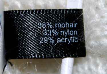 Close-up of black fabric tag showing composition of 38% mohair, 33% nylon, 29% acrylic in woman's white cowlneck mohair sweater.