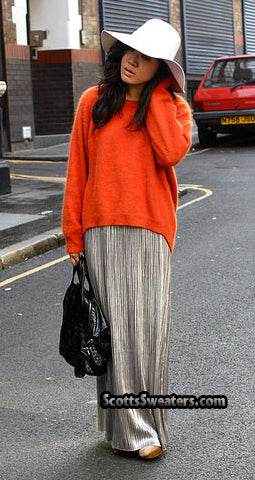 Woman wearing orange jewel-neck angora sweater and pleated metallic skirt with white wide-brim hat on city street