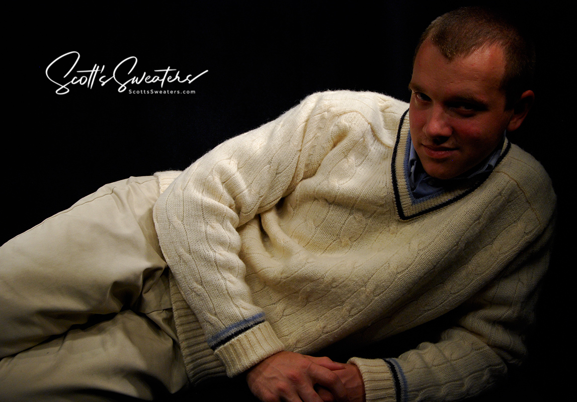 Man wearing retro classic ivory wool cable-knit tennis sweater with navy and French blue striped trim reclining against a dark background