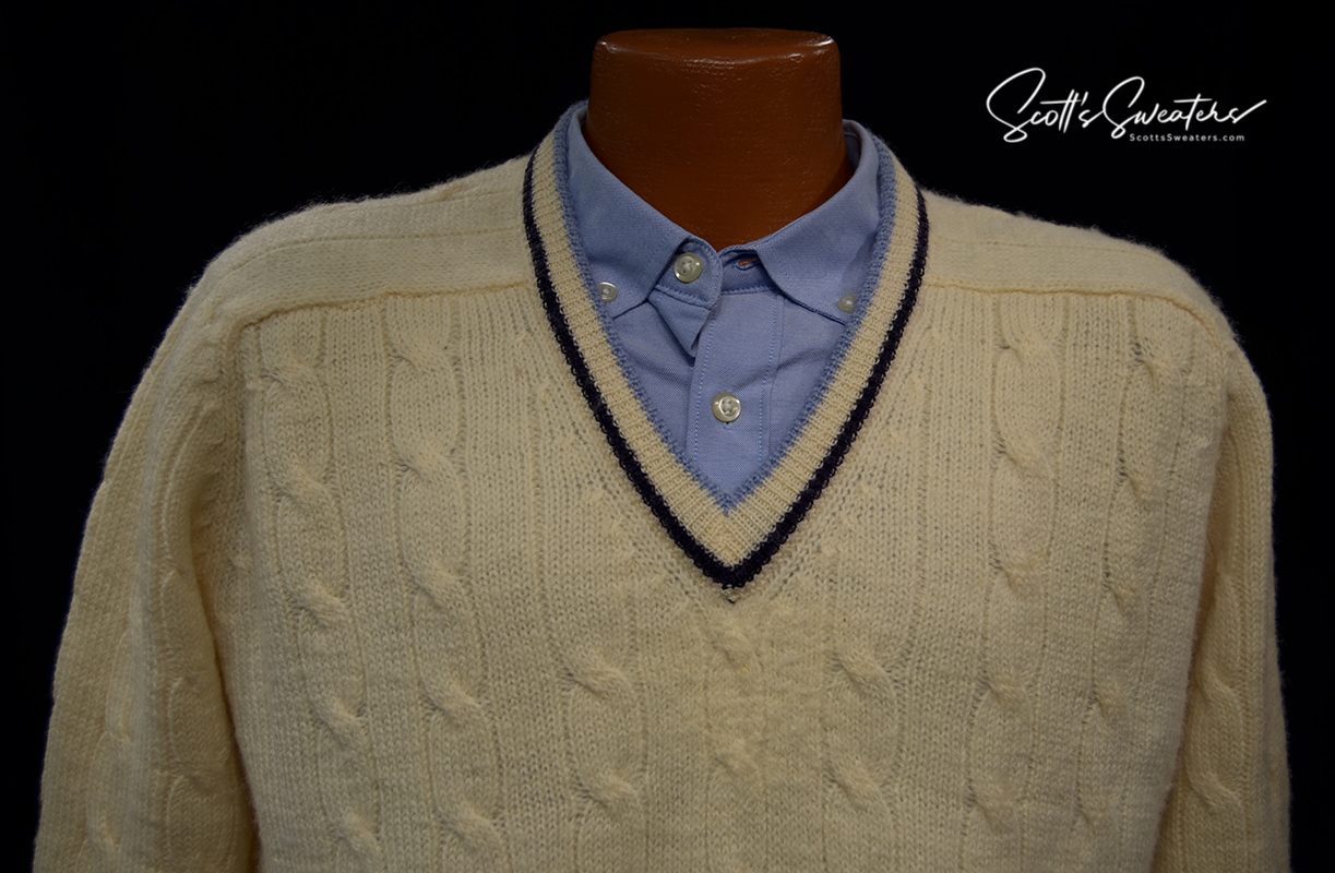 Ivory cable-knit wool tennis sweater with navy and French blue striped V-neck trim over blue button-down shirt