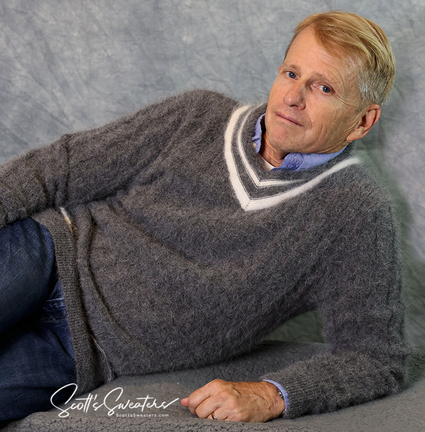 Man wearing a grey angora cableknit men's tennis sweater with white and dark grey stripes on the collar reclining against a textured grey background