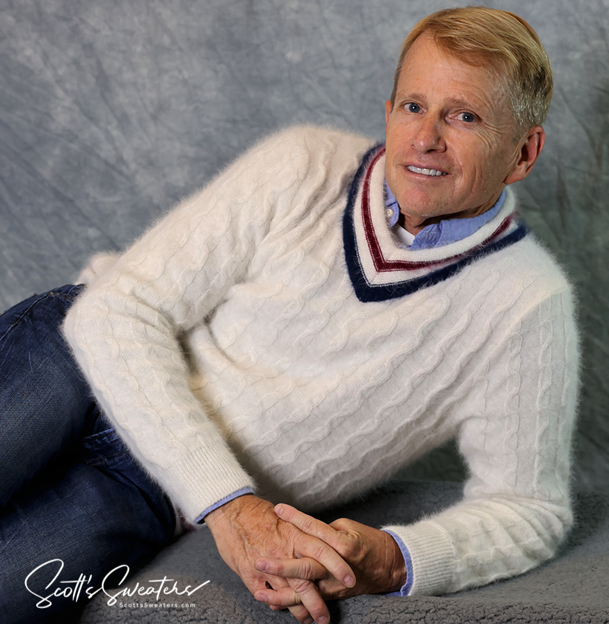 Man wearing a white cableknit angora men's tennis sweater with navy and red trim, reclining against a gray backdrop