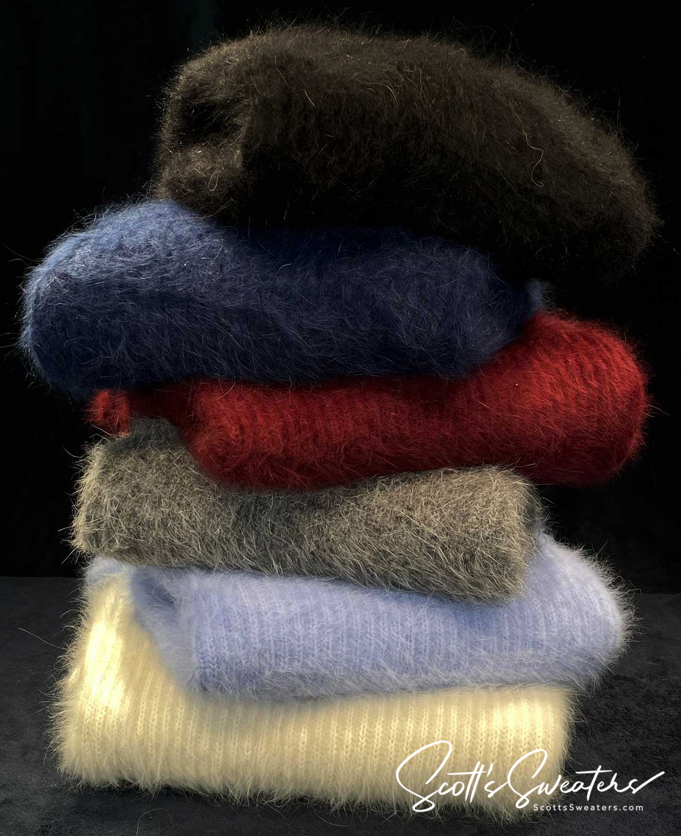 Stack of men's soft luxurious angora sweaters in assorted colors including black, navy, red, gray, light blue, and cream against a black background
