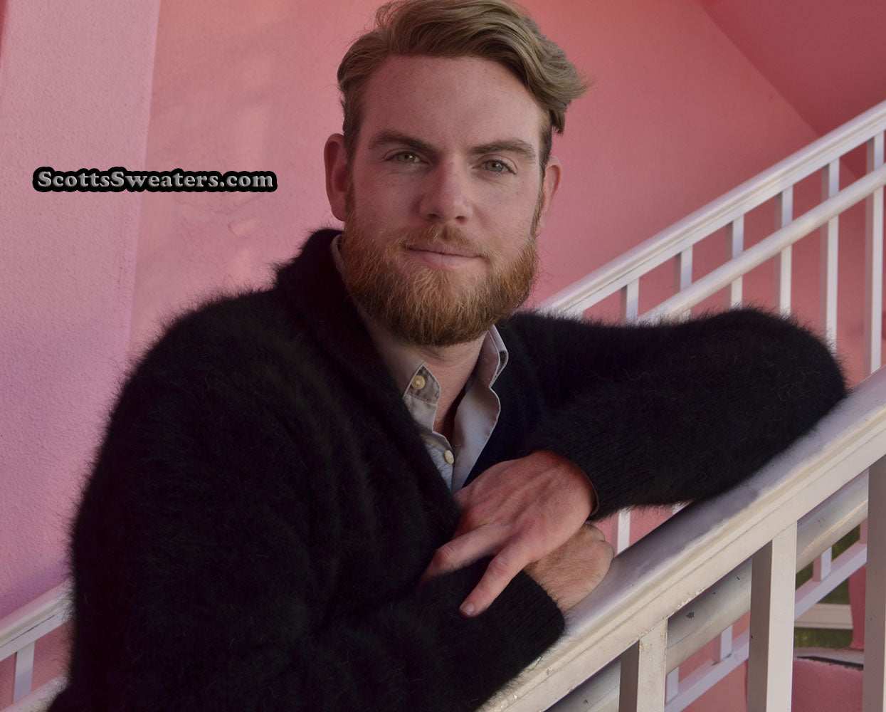 Man wearing a black men's solid-color angora shawl-neck cardigan sweater leaning on a white staircase railing.