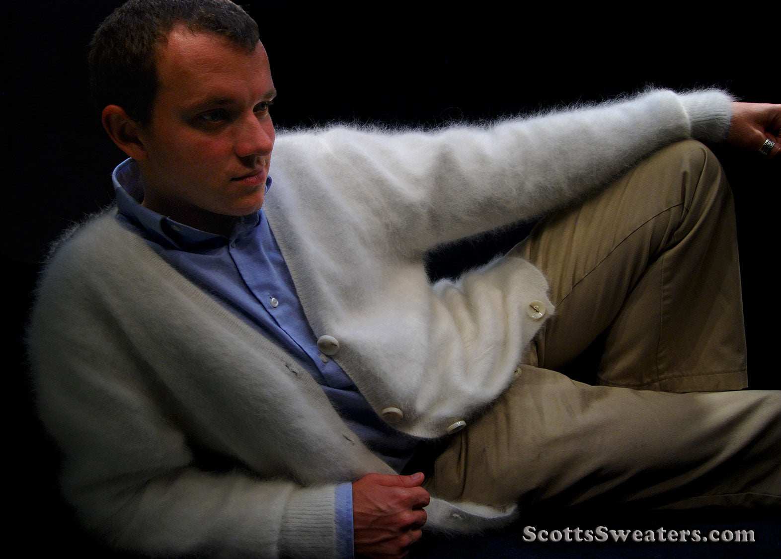 Man wearing men's ultra-soft angora white cardigan sweater with blue shirt and beige pants against black background