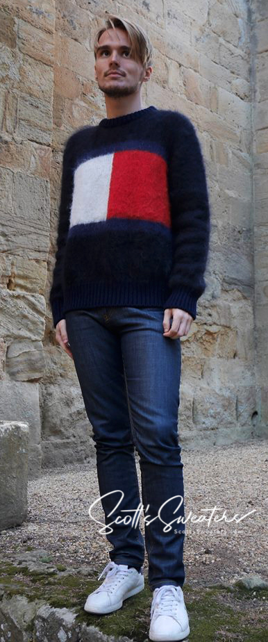 Men's black Tommy Hilfiger mohair crewneck sweater with large flag logo across chest, paired with dark jeans and white sneakers.