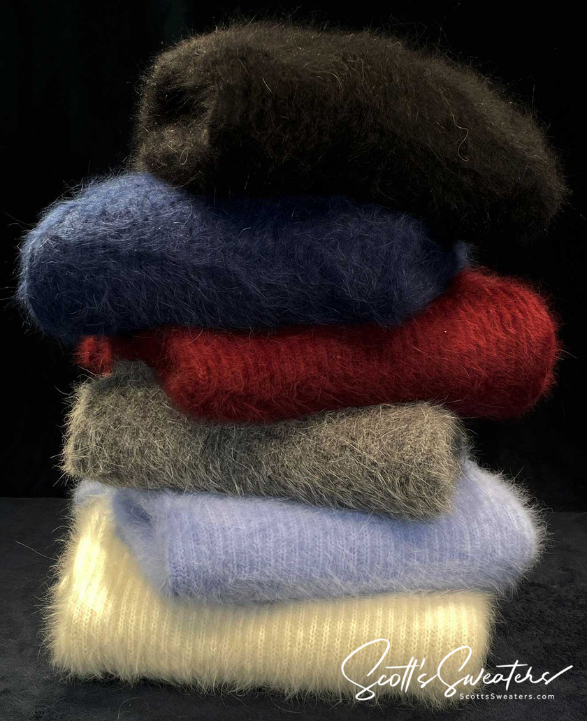 Stack of men's soft luxurious angora sweaters in assorted colors including black, navy, red, gray, light blue, and cream against a black background